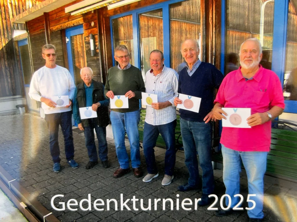Seven men hold medal certificates for Gedenkturnier 2025, posing outside a wooden building.